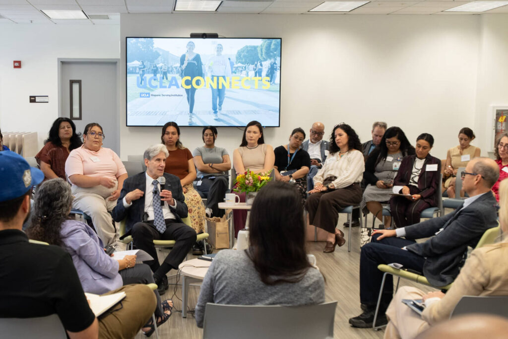 UCLA Chancellor Julio Frenk sits in the middle of a circle surrounded by a group of professionally dressed people. The screen behind him reads UCLA Connects