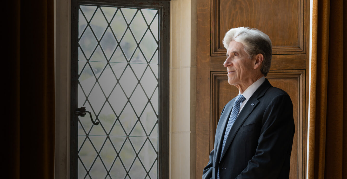 Chancellor Julio Frenk stands in profile by a glass window inside a wood-paneled UCLA building, looking out thoughtfully.