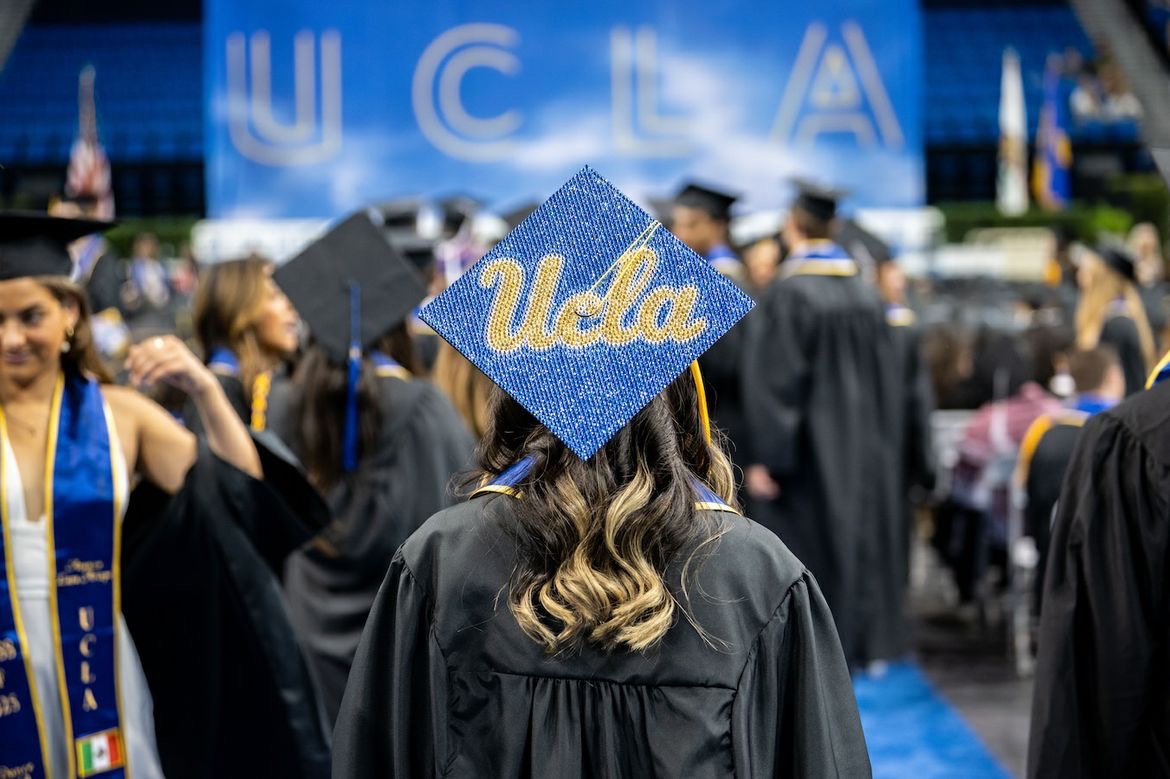 Woman with "UCLA" mortarboard, back turned, stands in front of a big "UCLA" sign in Pauley Pavilion