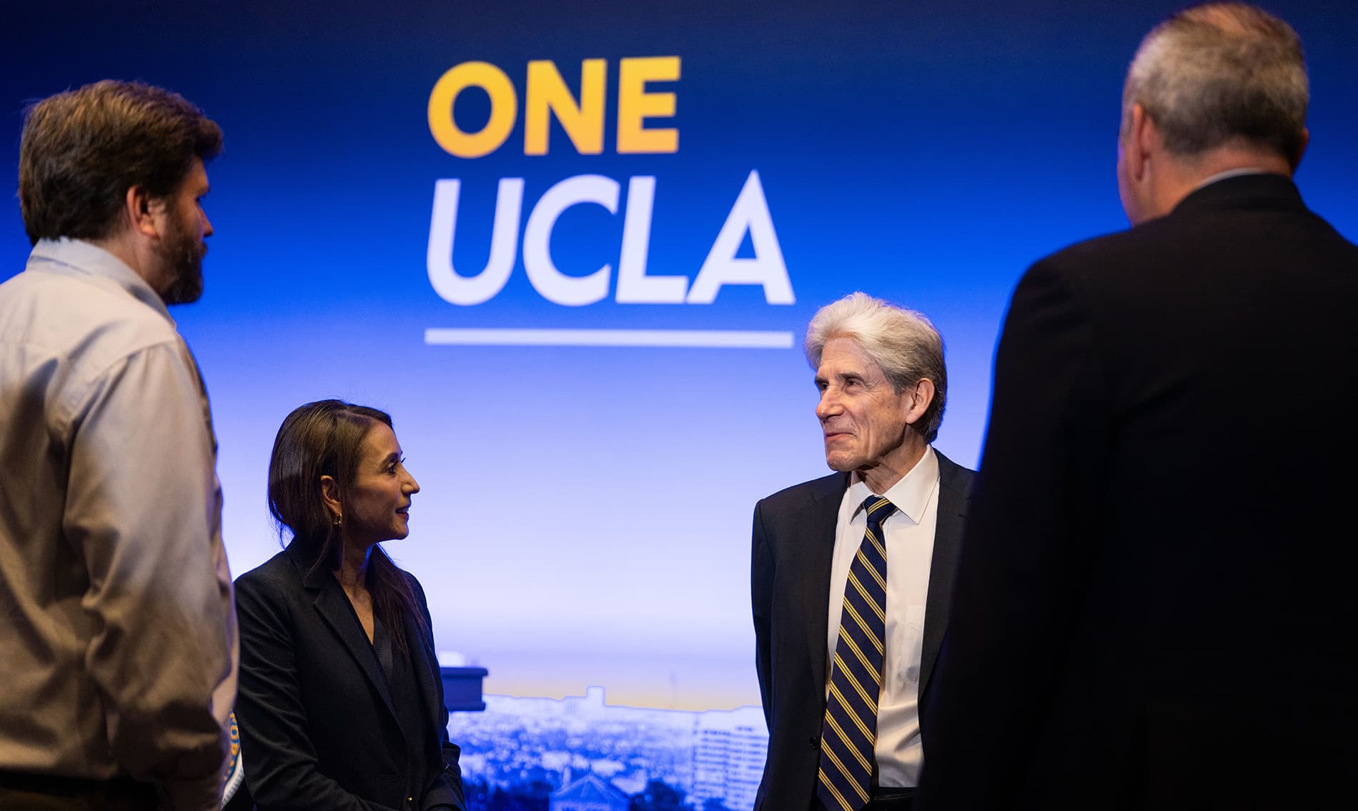 UCLA Chancellor Julio Frenk speaking with colleagues in front of a One UCLA backdrop.