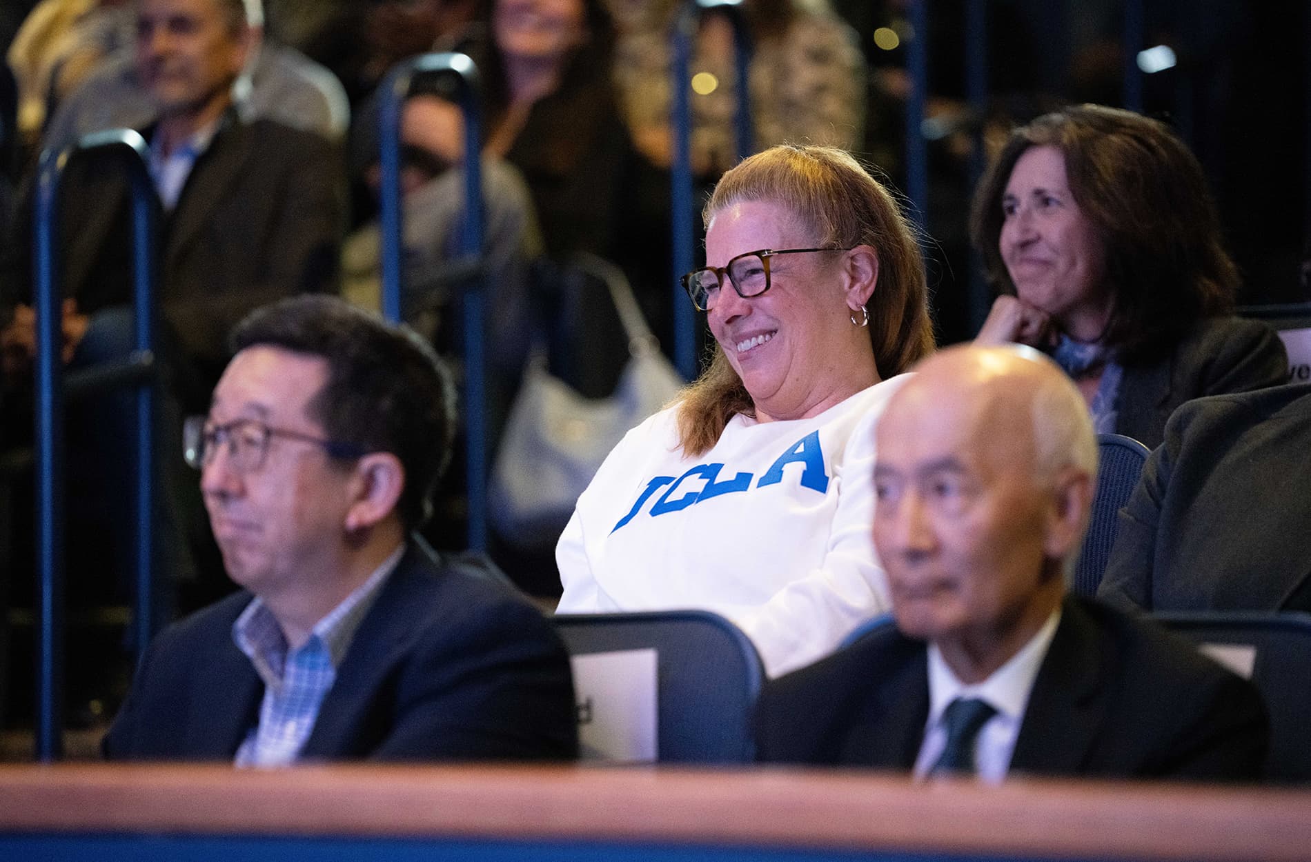 A woman wearing glasses and a white UCLA sweatshirt smiles while sitting in an audience during an event. Other attendees are visible around her in a darkened auditorium setting.