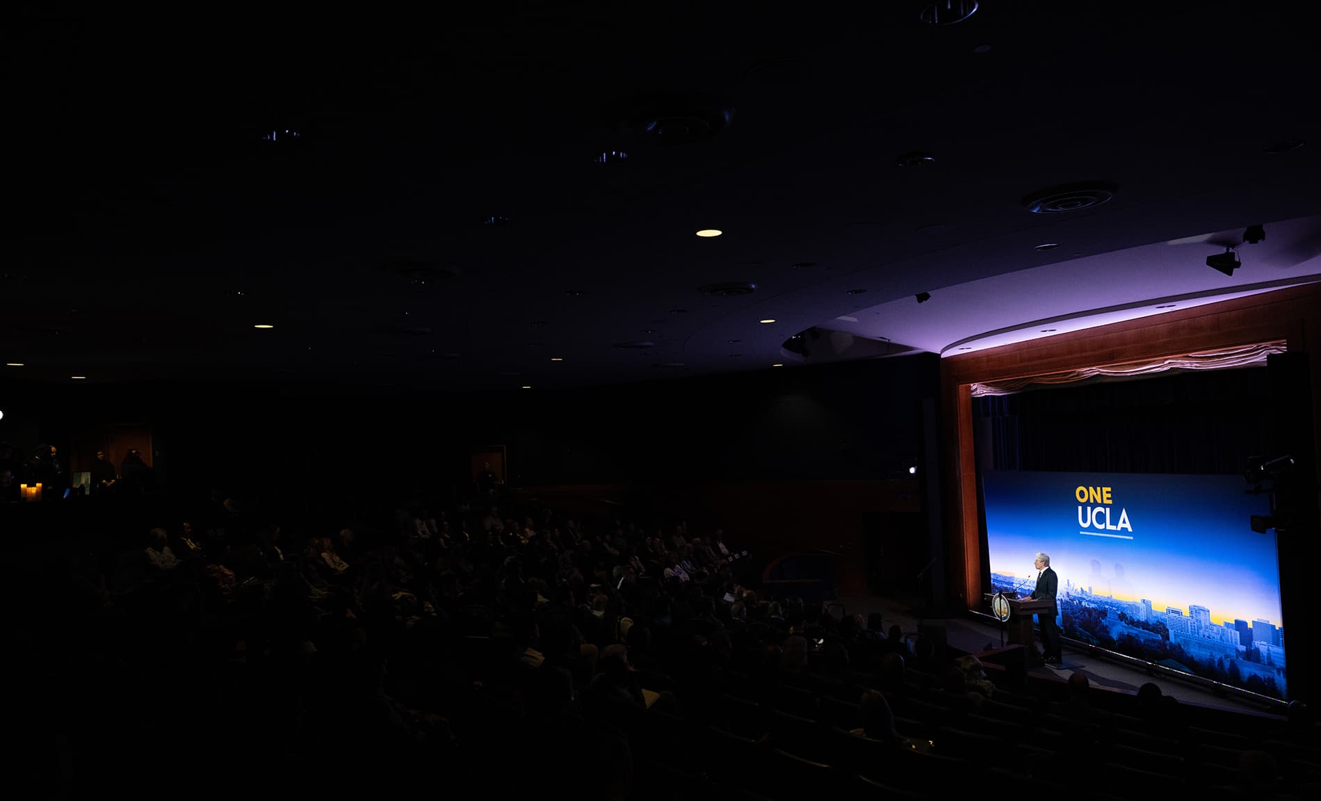 A wide shot from the back of a theater showing the silhouettes of an audience watching UCLA Chancellor Julio Frenk speak on stage.