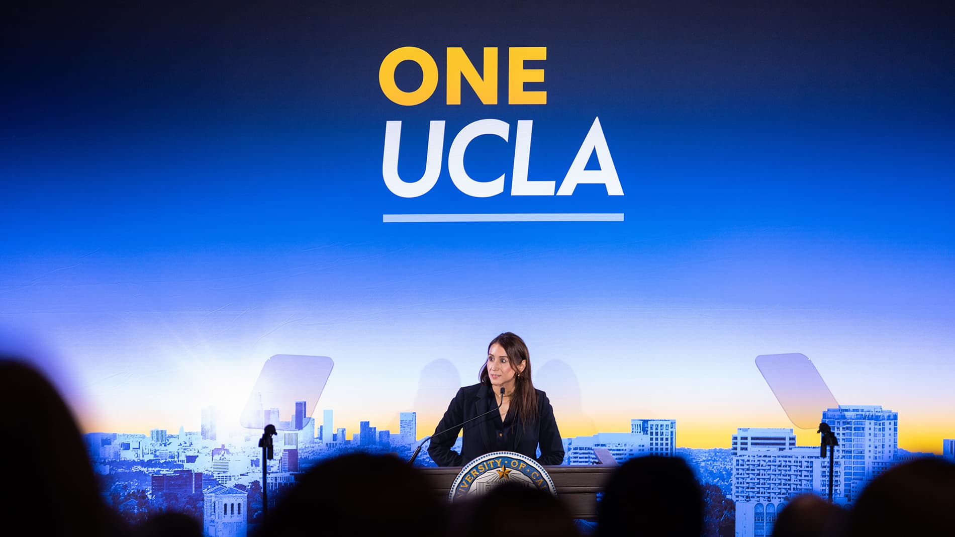 Adriana Galvan speaks at a podium in front of a large blue backdrop featuring the One UCLA logo, with a cityscape visible in the background.