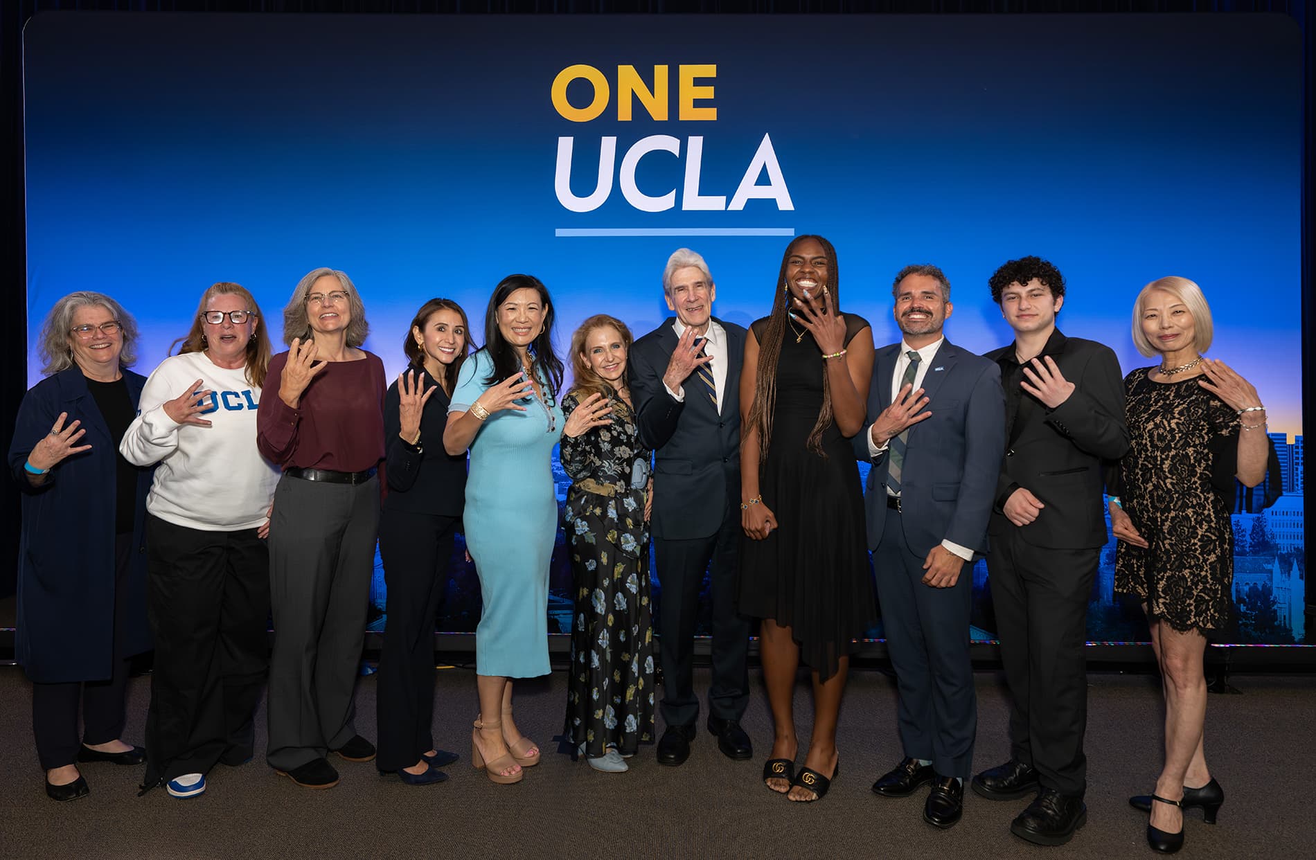 UCLA Chancellor Julio Frenk standing with a group of people holding up four fingers in front of a One UCLA backdrop.
