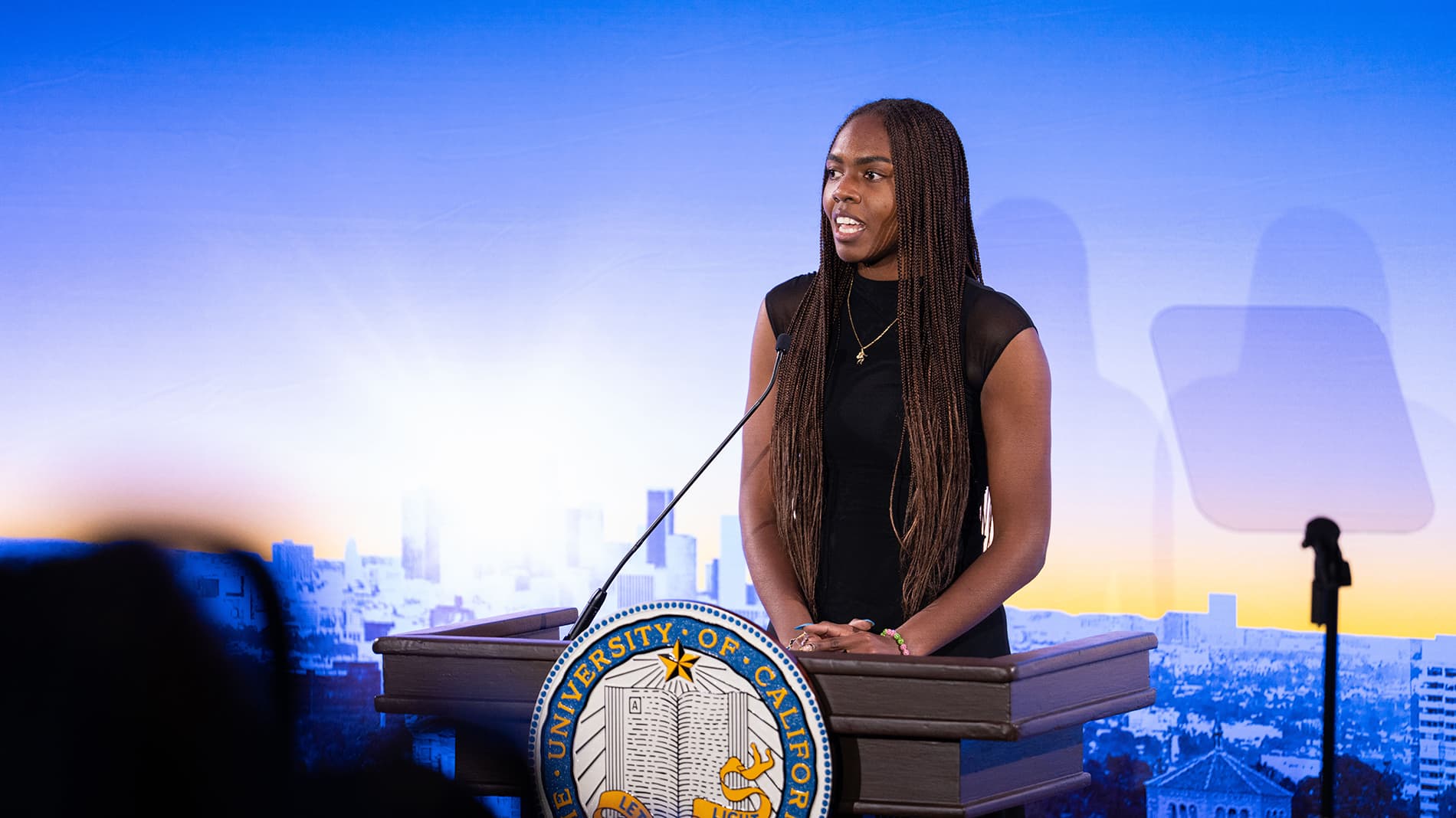 Naomi Hammonds stands at a podium bearing the University of California seal, speaking in front of a backdrop of a city skyline at dusk.