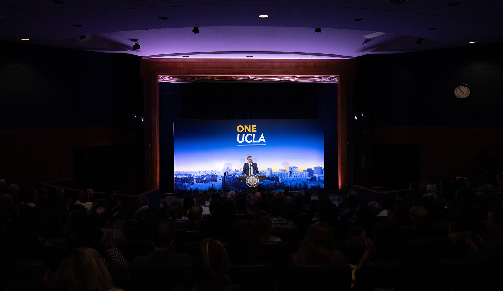 A wide shot from the back of a theater showing the silhouettes of an audience watching UCLA Chancellor Julio Frenk speak on stage.