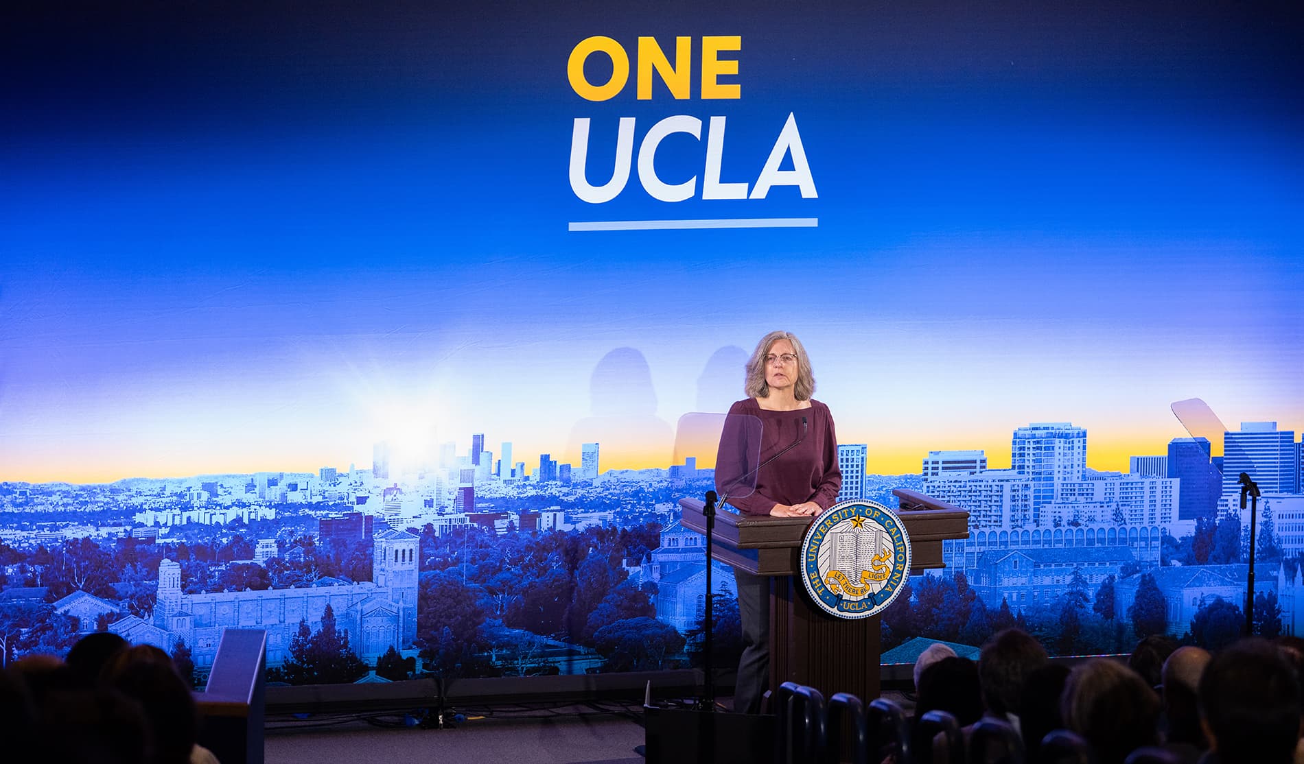 Andrea Ghez addresses an audience from a podium bearing the University of California seal, set against a large One UCLA backdrop and a cityscape at dawn.