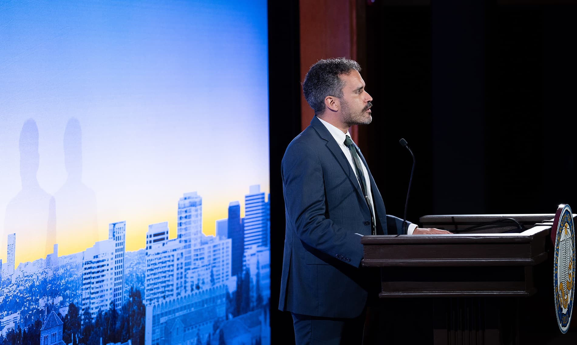 A profile shot of Michael McCormack standing at a lectern with a microphone. Behind him is a large, vibrant backdrop of a city skyline under a blue and yellow gradient sky, with the University of California seal visible on the front of the podium.